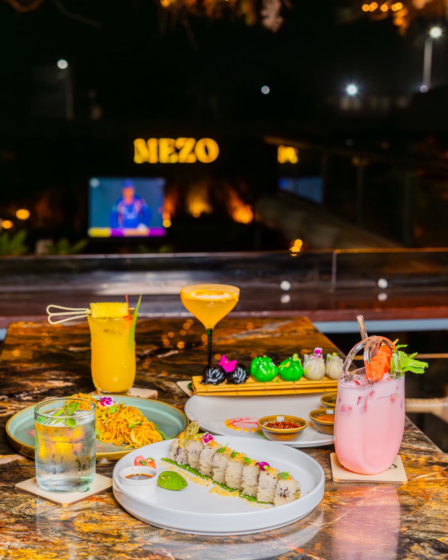 The glowing MEZO sign at night behind a table of cocktails and sushi